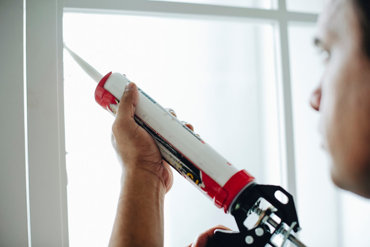 A man using a caulking gun to seal openings near his window. 